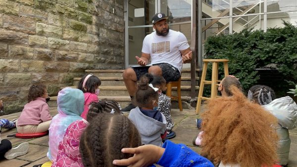 A man gestures to a group of children sitting before him.