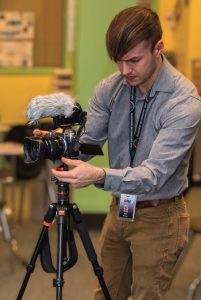 A man in a grey shirt and tan pants grabs a camera with a fluffy microphone sitting on a tripod.