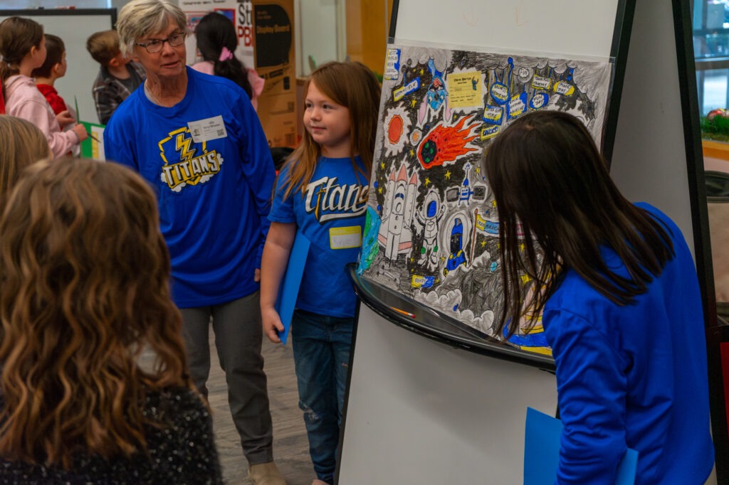 Two elementary students stand alongside a display focused on PBIS efforts while speaking with a group of students and teachers.